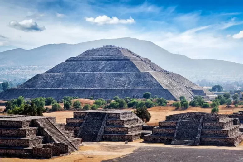 Vista panorámica de la Pirámide del Sol en Teotihuacán, México, rodeada de estructuras menores y vegetación, con montañas al fondo y cielo despejado.